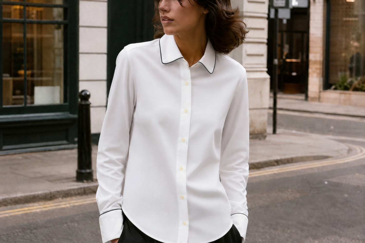 A woman wearing a white long-sleeve button-up shirt with black piping on the collar and cuffs, standing on a city street.